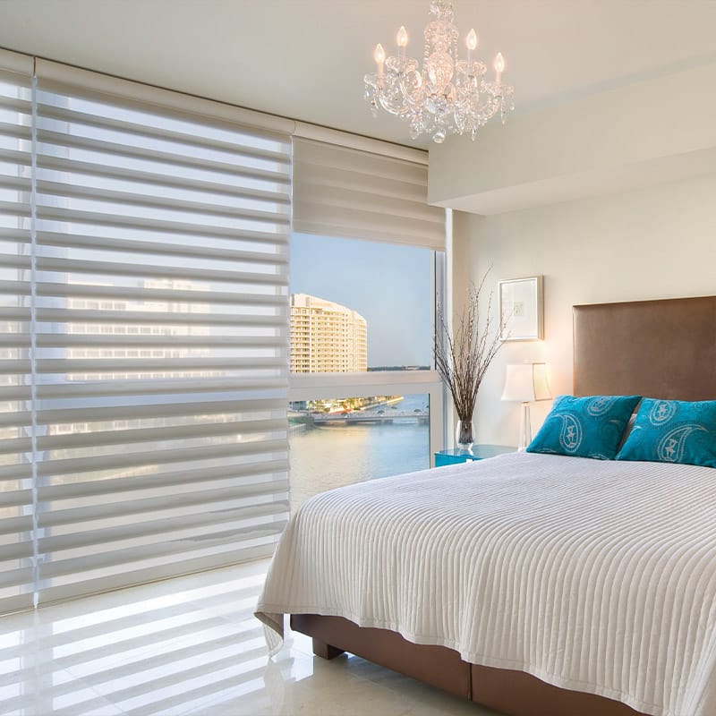 Sunny bedroom with large window overlooking a waterway and cityscape, cozy bed with blue pillows, elegant chandelier, and contemporary decor, ideal for relaxation and city living.