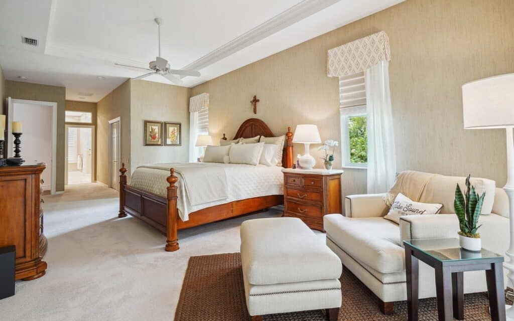 Expansive bedroom with beige walls, linen curtains, wooden bed frame, nightstands, and a sitting area with a sofa, ottoman, and decor plants, creating a warm, inviting retreat.