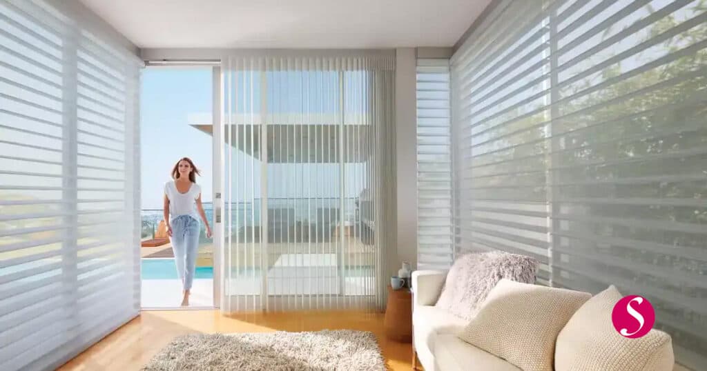 Smooth white horizontal blinds in a modern living room with a woman enjoying sunlight and outdoor views, showcasing sunshade window treatments by In The Shade for stylish, functional window coverings.