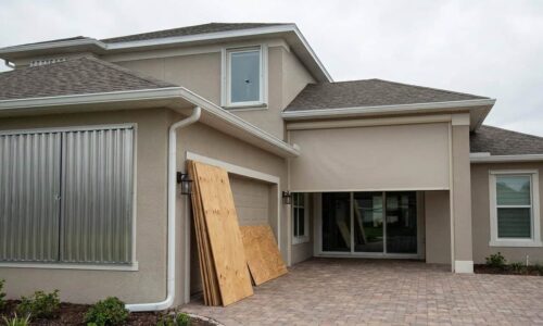 Modern house exterior with renovation work underway, plywood boards covering garage, brick driveway, and new window treatments visible.